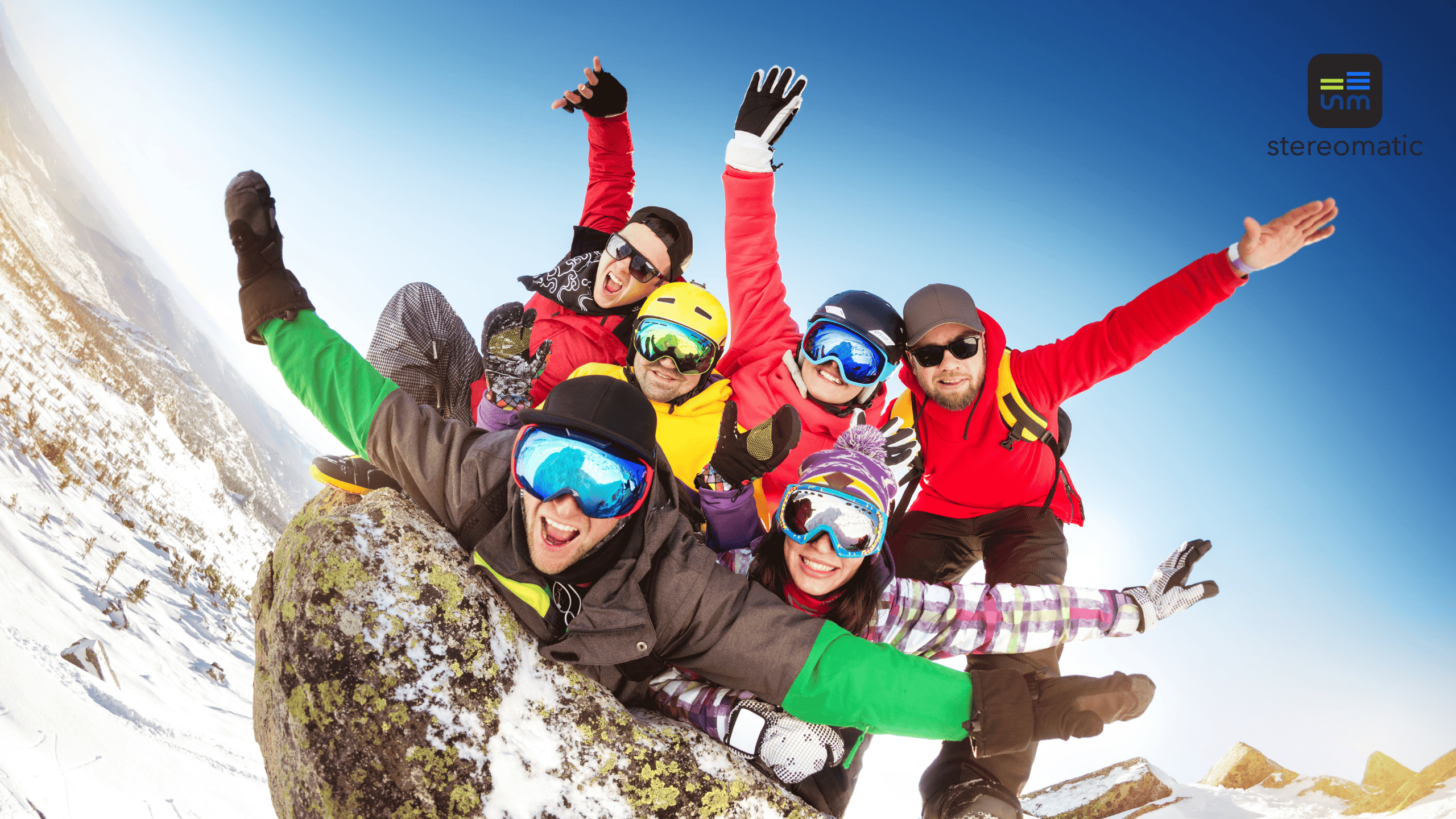 Grupa uśmiechniętych uczestników zawodów narciarskich pozuje wspólnie na szczycie ośnieżonej góry, unosząc ręce w geście zwycięstwa i integracji.